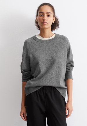 Suéter gris hecho de una tela suave con un escote redondo y puños de canalé. Presenta un corte holgado y aberturas laterales en el dobladillo. Combinado con pantalones negros.