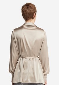 Beige satinblus med knytmidja, långa ärmar, sjalkrage och rynkade detaljer bak. Slät textur och lös, flytande design.