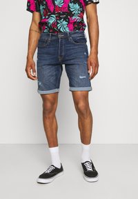 Denimshortsit, joissa on keskisininen sävy, kulutettuja yksityiskohtia ja käännetyt reunat. Yhdistetty rohkeaan, moniväriseen kukkapaitaan ja mustiin tennareihin.