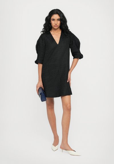 Zwarte jurk met een V-hals, gestructureerde stof en korte puff mouwen. Gecombineerd met een blauwe clutch en witte puntige flats.