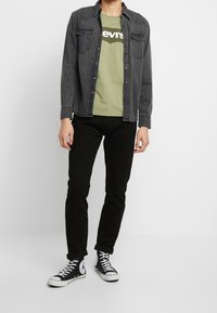 Camicia di jeans nera sopra una t-shirt verde con logo, abbinata a jeans neri e sneakers alte nere con un design grafico.