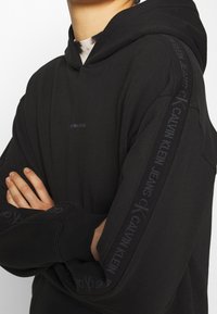 Schwarzer Kapuzenpullover aus weichem Material. Mit Logo entlang der Ärmel und einem minimalistischen Design auf der Vorderseite. Rippbündchen an den Ärmeln und am Saum.