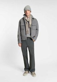 Grau melierte Wolljacke mit zwei vorderen Taschen, getragen über einem hellgrauen Pullover, dunkelgrauen Hosen und einer passenden Strickmütze.