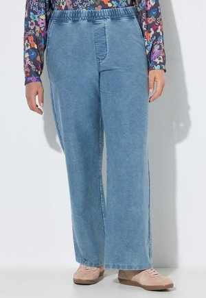 Person trägt hellblaue, weite Hosen, ein buntes, langärmeliges, geblümtes Oberteil und beigefarbene Schnürschuhe und steht an einer hellen Wand.