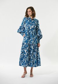 Blaues Blumen-Maxikleid mit knöpfbarem Kragen, langen Puffärmel, gestuften Design und einem leichten Stoffmuster in verschiedenen Blau- und Weißtönen.