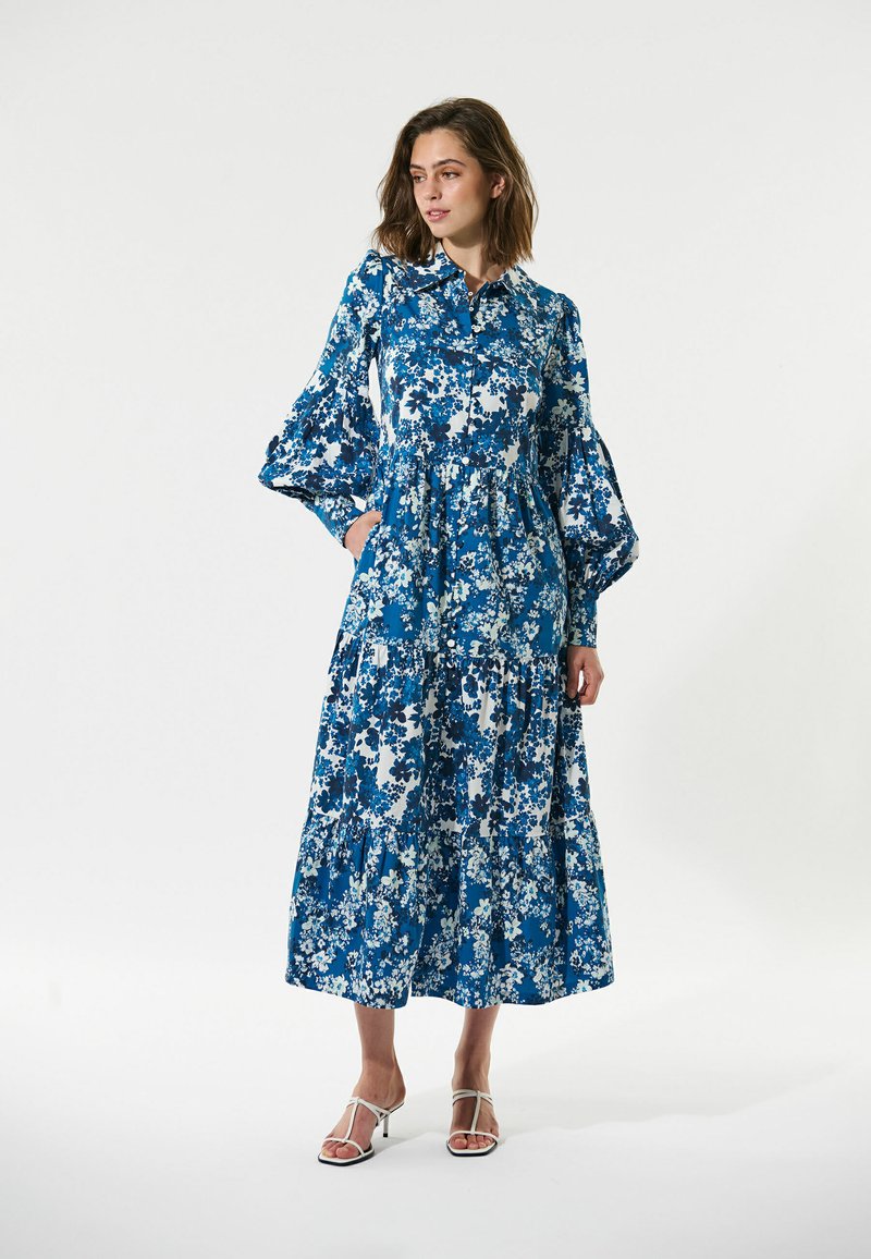 Blaues Blumen-Maxikleid mit knöpfbarem Kragen, langen Puffärmel, gestuften Design und einem leichten Stoffmuster in verschiedenen Blau- und Weißtönen.