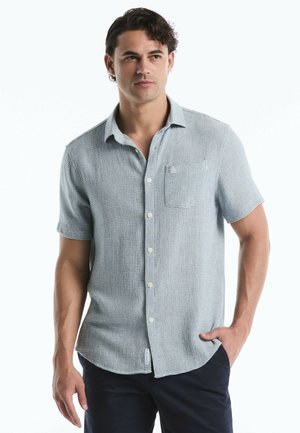 Man draagt lichtblauw overhemd met korte mouwen en donkere broek, staat met één hand in de zak en kijkt lichtjes opzij.