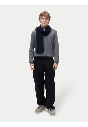 Junger Mann trägt grauen Pullover, schwarzen Schal, lockere schwarze Hose und beige Schuhe, steht vor schlichtem hellem Hintergrund.