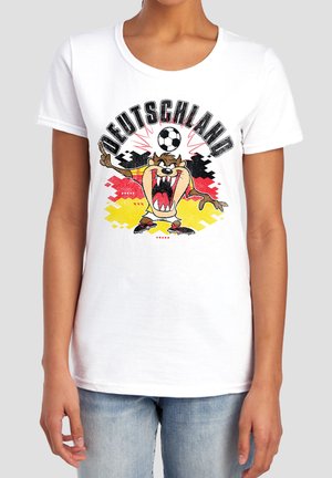 Person trägt ein weißes T-Shirt mit einem Cartoon-Tasmanischen Teufel, der einen Fußball balanciert, und "Deutschland" Text vor einem schwarzen, roten und gelben Hintergrund.