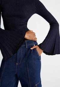 Personne portant un haut bleu foncé côtelé à manches longues avec des poignets évasés et un jean en denim bleu taille haute, mains dans les poches.