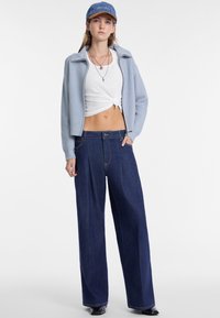 Giacca azzurro chiaro a costine con zip sopra una maglietta bianca cropped, abbinata a jeans larghi in denim scuro e un cappellino blu. Accenti di gioielli semplici.