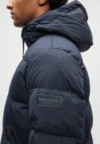 Casaco acolchoado azul-marinho com capuz, textura matelassê costurada, bolso lateral com fecho de correr e etiqueta de marca em destaque na manga.