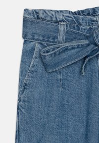 Světle modré džíny s elastickým pasem na šňůrku, které mají detail vázané mašle a přední kapsy. Tkanina má jemnou strukturu.