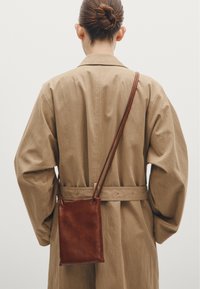 Brązowa skórzana torba na ramię, prostokątna, gładka w dotyku, wisząca na cienkim pasku na beżowym płaszczu z szerokimi rękawami.