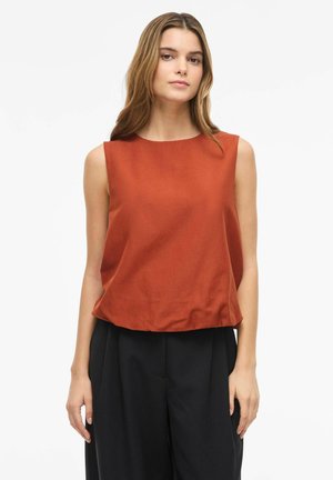 Woman wearing a sleeveless rust orange top and black wide-leg pants standing against a plain white background.