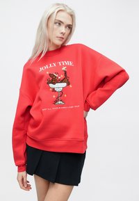 Κόκκινο υπερμεγέθες φούτερ με κέντημα μιας γιορτινής σκηνής, που περιλαμβάνει το κείμενο "ΧΑΡΟΥΜΕΝΟΣ ΧΡΟΝΟΣ" και "ΕΥΧΟΜΑΙ ΟΛΕΣ ΣΟΥ ΟΙ ΕΥΧΕΣ ΝΑ ΓΙΝΟΥΝ ΠΡΑΓΜΑΤΙΚΟΤΗΤΑ." Μείγμα βαμβακιού.