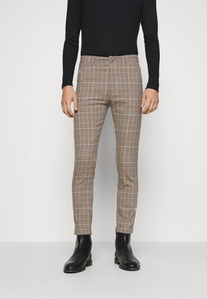 Person trägt schlanke, beige karierte Hose, schwarzes Langarmshirt und schwarze Stiefeletten und steht vor einem einfarbigen Hintergrund.