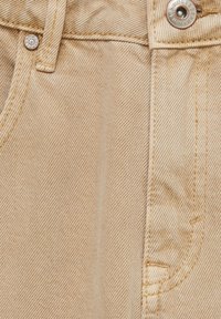 Tissu en denim beige en gros plan montrant une boucle de ceinture, le bord de la poche, les coutures et des rivets métalliques avec le texte de la marque.