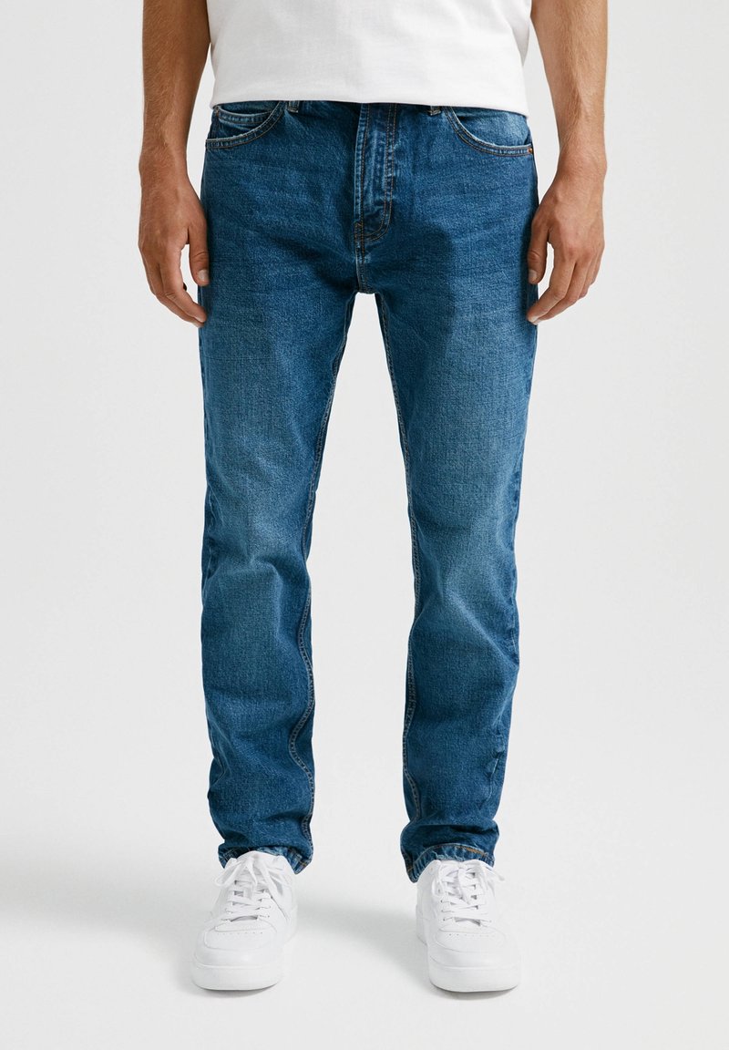 Vaqueros de mezclilla azul claro con corte recto, cinco bolsillos y un desgaste sutil, combinados con zapatillas blancas.