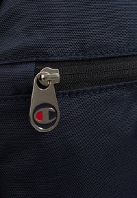 Granatowa zasuwana kieszeń z materiału, z teksturowanym wzorem i srebrnym suwakiem z logo w czerwonych i granatowych okrągłych akcentach.