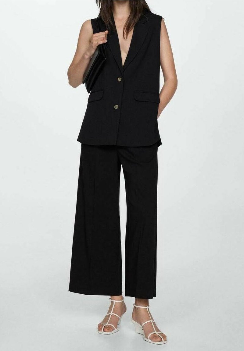 Femme portant un blazer noir sans manches et un pantalon noir à jambes larges, tenant un sac noir, associé à des sandales compensées blanches à brides.