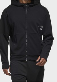 Svart munkjacka med dragkedja tillverkad av texturerad tyg, med en frontficka, huva med dragsko och subtila logodetaljer.