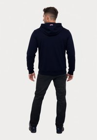 Bărbatul stă cu spatele, purtând un hanorac bleumarin cu gluga lăsată, pantaloni negri și pantofi negri, pe un fundal alb simplu.