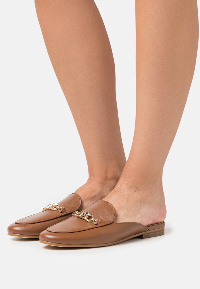 Pieds portant des mocassins marron en cuir sans talon avec un détail de chaîne dorée, affichés sur un fond clair uni.