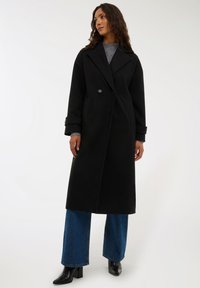Czarny wełniany płaszcz oversize z podwójnym zapięciem, dużymi klapami i detalami w postaci guzików. Zestawiony z niebieskimi spodniami o szerokich nogawkach i czarnymi botkami.