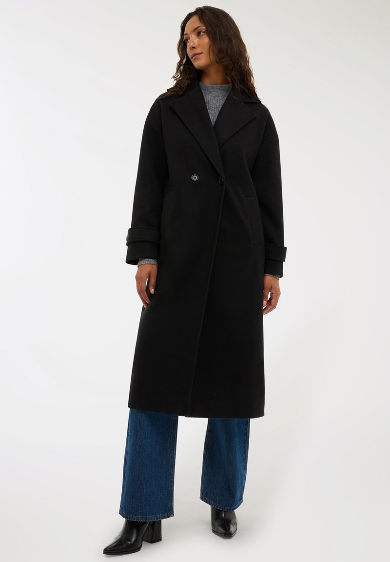 Czarny wełniany płaszcz oversize z podwójnym zapięciem, dużymi klapami i detalami w postaci guzików. Zestawiony z niebieskimi spodniami o szerokich nogawkach i czarnymi botkami.