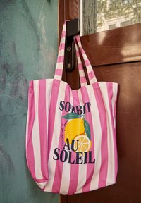 Sac fourre-tout rayé rose et blanc avec image de citron et texte suspendu à une poignée de porte marron contre un mur vert.