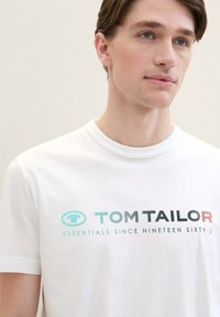 Bílá bavlněná tričko s kulatým výstřihem, s logem "TOM TAILOR" ve tyrkysové a černé barvě s červeným akcentem, hladká textura.