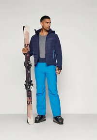 Ski outfit featuring a navy padded jacket, gray shirt, bright blue pants, and black ski boots. Holding a ski with black bindings.
