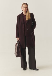 Burgundský vlnený kabát s vykrojenými klopami a dvěma knoflíky, doplněný ladící košilí a širokými černými kalhotami, nesoucí hnědou koženou tašku.