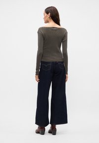 Γυναίκα που στέκεται με την πλάτη στραμμένη, φοράει ένα ελαιόχρωμο τοπ με ανοιχτούς ώμους και μακριά μανίκια, φαρδιά μπλέ τζιν και καφέ ψηλοτάκουνες μπότες.