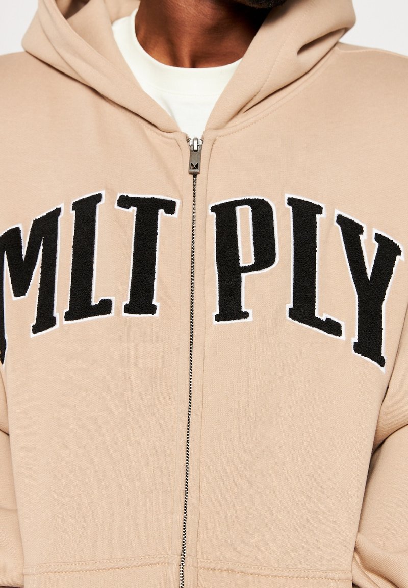 Personne portant un hoodie beige à fermeture éclair avec de grandes lettres noires MLTPLY sur la poitrine, t-shirt blanc partiellement visible en dessous.