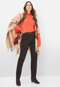 Donna che indossa un maglione arancione a collo alto, uno scialle fringed a righe, pantaloni neri e stivali alla caviglia marrone scuro, in piedi su uno sfondo bianco.
