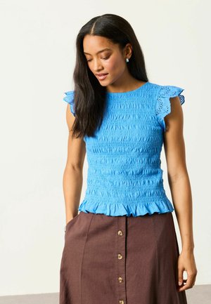 Young woman with long dark hair wearing a blue textured sleeveless top with ruffled edges and a brown buttoned skirt, hands in pockets.