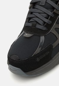 Melnas sporta kurpes ar tekstūru auduma augšdaļu un gludām akcentiem, pelēkiem zoleiem un atstarotājiem. Malā ir iekļauts Gore-Tex logo.