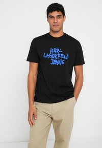 Juodas medvilninis T-shirt, puikuojantis mėlynai purškiamu tekstu "KARL LAGERFELD JEANS". Trumpi rankoviai, apvalus kaklas, laisvas kirpimas.