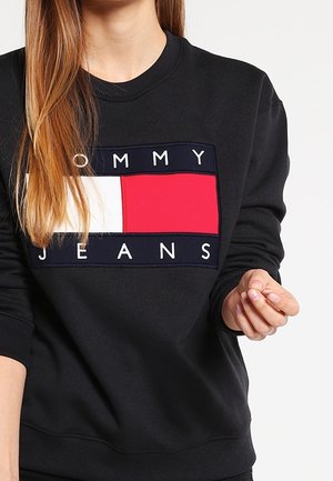 Osoba nosząca czarną bluzę z czerwono-biało-granatowym logo Tommy Jeans oraz z długimi jasnobrązowymi włosami częściowo widocznymi.