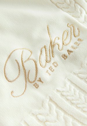 Bela pletiva svijetle boje s reljefnim kabelnim uzorkom i zlatnim izvezenim tekstom "Baker by Ted Baker." Mekana tekstura, čist dizajn.
