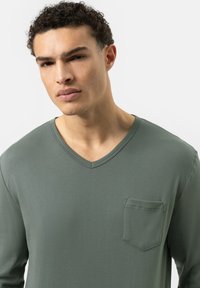 Jonge man met kort krullend haar, gekleed in een groene V-hals lange mouw shirt met een borstzakje, die iets opzij kijkt.