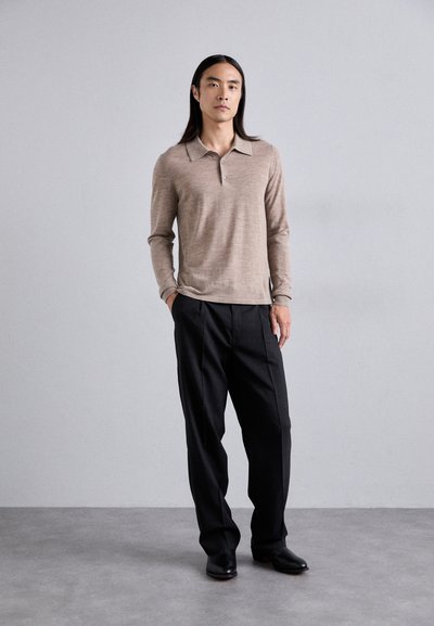 Polo shirt beige a maniche lunghe con colletto e patta a tre bottoni, abbinato a pantaloni neri a gamba larga e scarpe eleganti nere.
