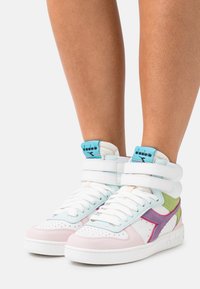 Vita hög-top sneakers med pastellrosa, lila, gröna accentfärger, vita snören och en turkos Diadora-logotyp på tungan, bärs på bara ben.