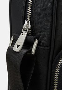 Fermeture éclaire d'un sac à dos en nylon noir avec surface texturée, comportant un tirette métallique et une sangle ajustable. Gros plan sur les détails du matériel.