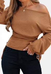 Brązowy, prążkowany top z odkrytymi ramionami i długimi, rozkloszowanymi rękawami; szczegóły z teksturowanej tkaniny; sparowany z czarnymi dżinsami i warstwowymi pierścionkami na dłoni.