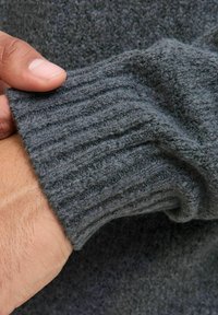 Grauer, gestrickter Pulloverbund mit einem gerippten Muster aus weichem, flauschigem Material. Eine Hand hält vorsichtig den Rand.