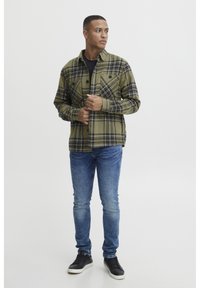 Grünes kariertes Hemd mit schwarzen Akzenten, durchgehende Knopfleiste, zwei Brusttaschen, über einem schwarzen T-Shirt getragen, kombiniert mit blauen Skinny-Jeans und schwarzen Sneakers.