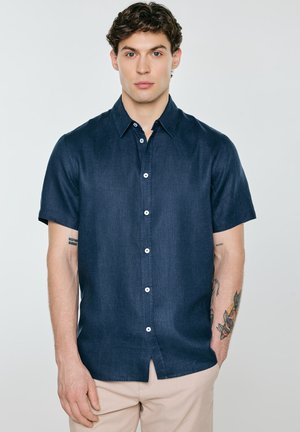 Młody mężczyzna z krótkimi kręconymi włosami, ubrany w ciemnoniebieską krótką koszulę zapinaną na guziki i beżowe spodnie, stoi na tle jednolitego jasnego tła.